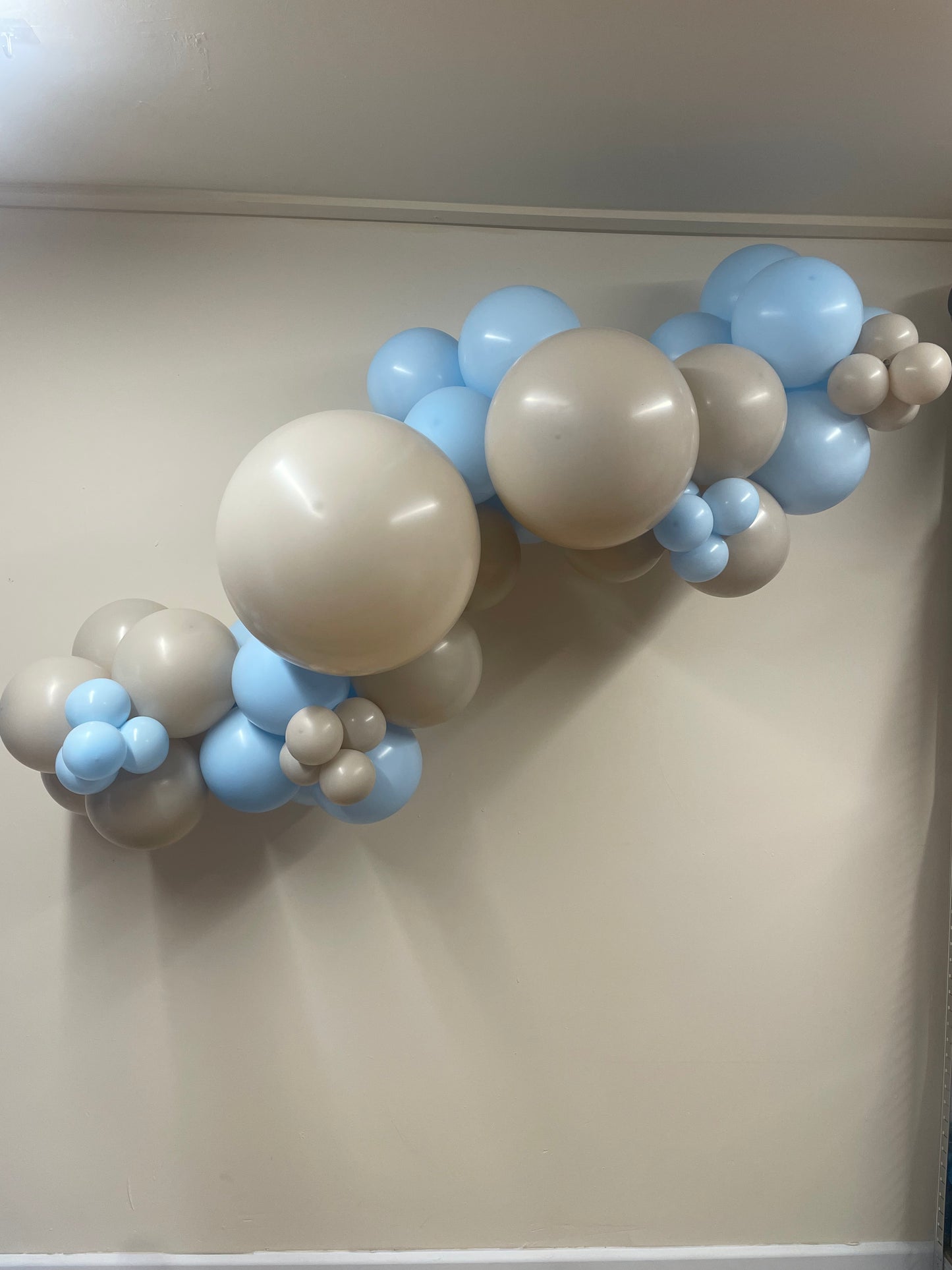 Balloon arch with beige, blue, and brown balloons against a plain wall.