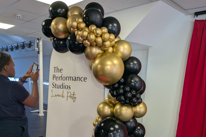Balloon arch with gold and black balloons in front of a branded backdrop at The Performance Studios.