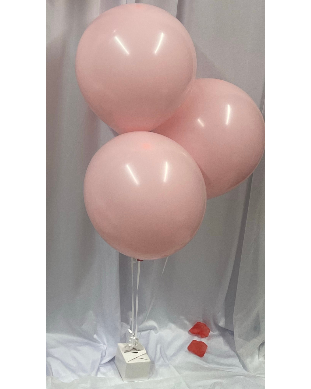 Three pink balloons tied to a small white box with red hearts on a light gray background