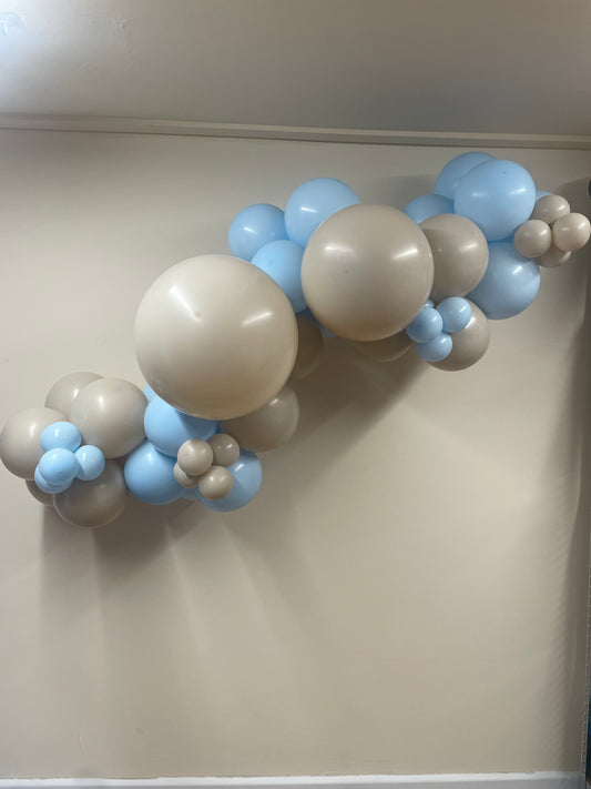Balloon arch with beige, blue, and brown balloons against a plain wall.